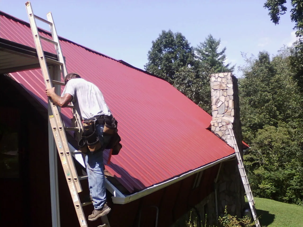 Roofer on a ladder beside a red metal roof and stone chimney — Soffit & Fascia Repair by Roofing Brothers in Girard